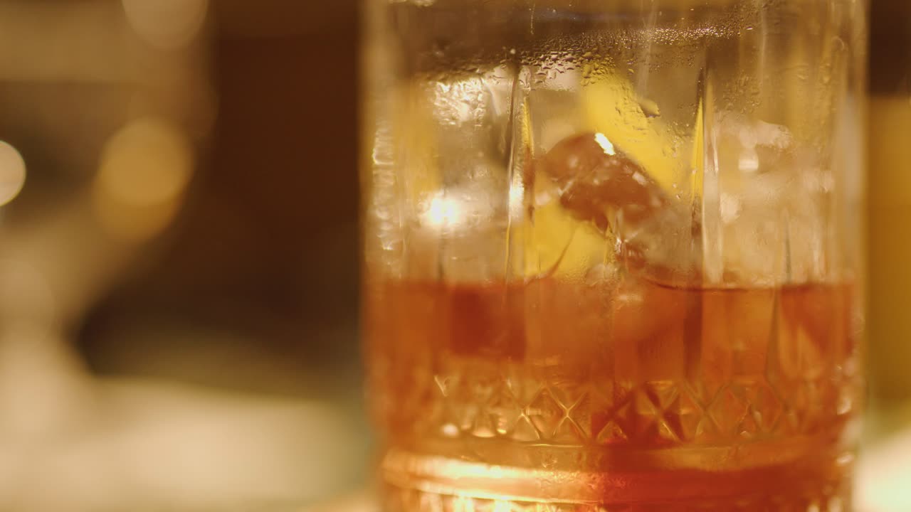A hand grasps a chilled Negroni cocktail with ice and orange peel garnish on a bar counter, under warm, moody evening lighting with shallow focus