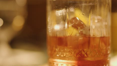 a hand grasps a chilled negroni cocktail with ice and orange peel garnish on a bar counter, under warm, moody evening lighting with shallow focus