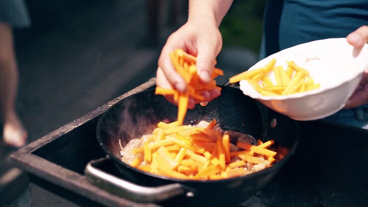 Close-up Young man makes pilaf in a cauldron on a grill on an open fire Add carrots to pork