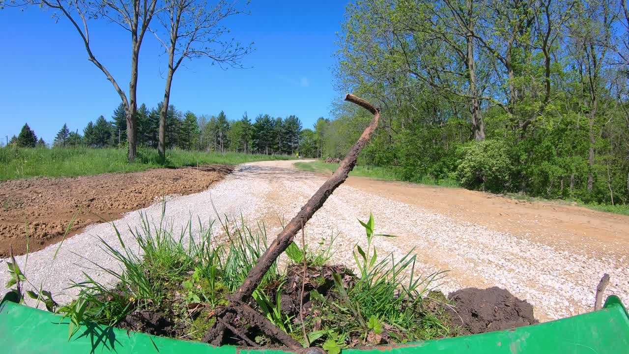 pov, operando un pequeño tractor verde y usando un cubo de cargador para mover tierra y escombros en un carril de grava recién pavimentado rodeado de un pasto y árboles