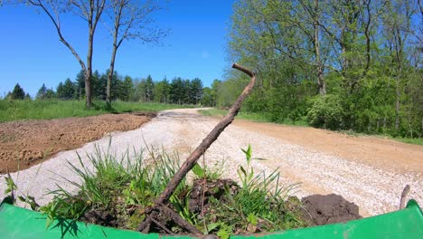 pov, operating small green tractor and its using loader bucket to move dirt and debris on newly graveled lane surrounded by a pasture and trees