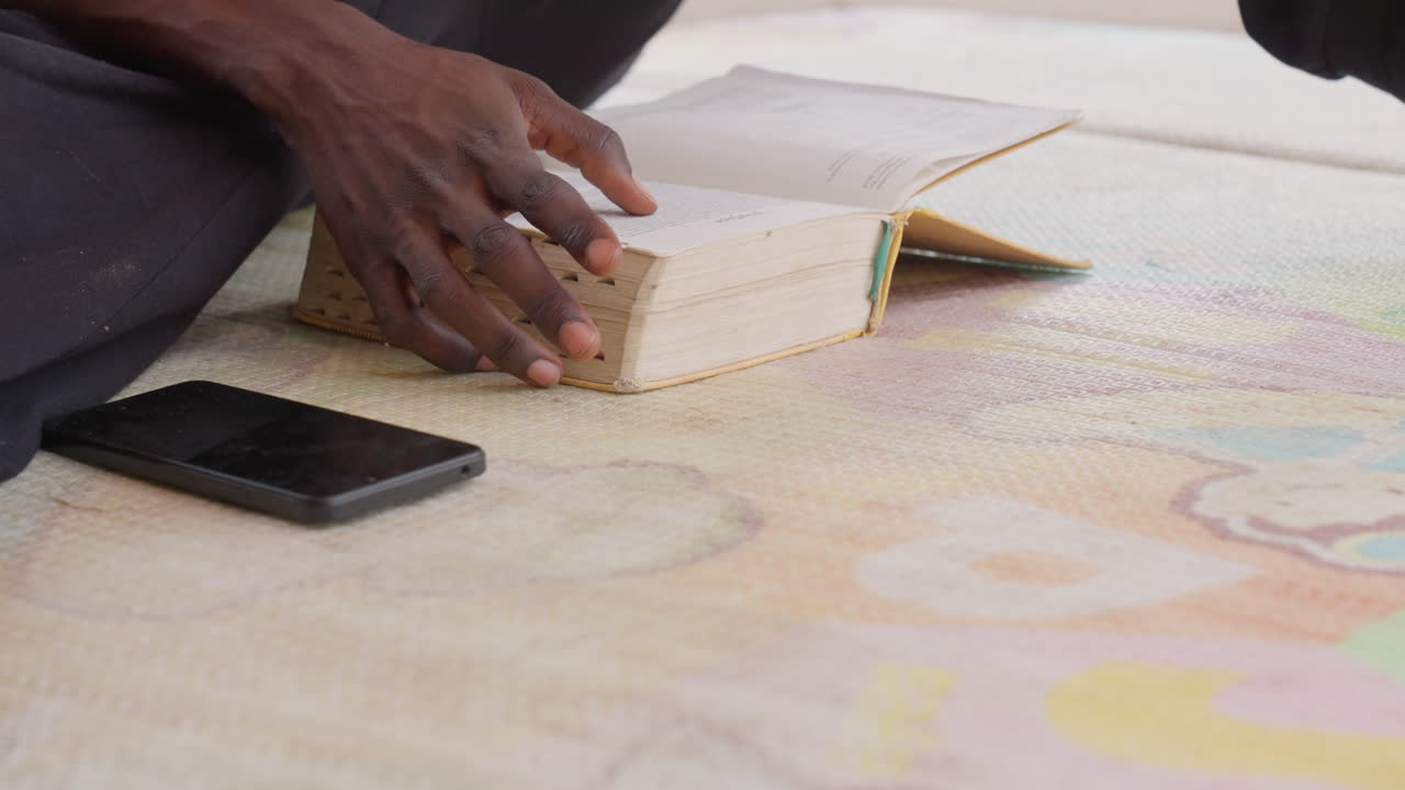 Black student hands on book phone, fingers tracing lines on pages, colorful patterned rug beneath, focused revision vibe, modern device at side, quiet indoor concentration, mindful study moment