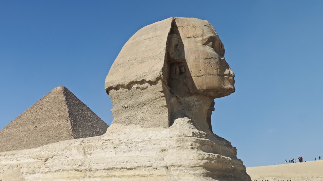 Great Sphinx of Giza, carved from limestone bedrock, faces right, with its head and body clearly visible against a bright blue sky. The Pyramid of Khufu rises in the background