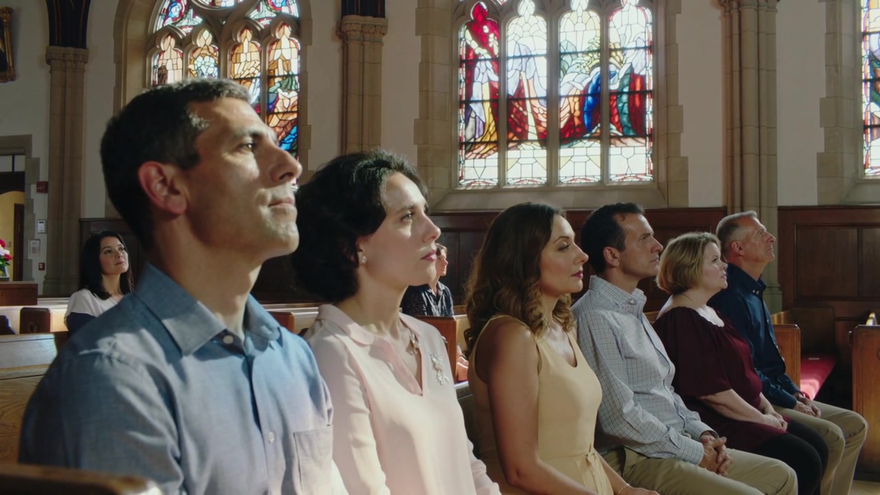 Seis adultos sentados movendo as cabeças no banco enquanto o culto começa na nave da igreja, com o brilho dos vitrais.