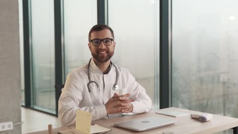 el médico varón diligente sentado en el escritorio con una computadora portátil le pide al paciente que entre a la oficina y se siente enfrente