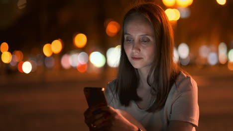 por la noche, una joven se sienta en la hierba de una gran ciudad y mira a la pantalla de su teléfono inteligente, escribe un mensaje y mantiene su blog personal.