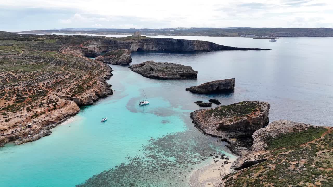 日中のマルタ島コミノにあるブルーラグーンの空撮。透明なターコイズブルーの海にはボートが停泊し、岩だらけの崖、小さな入り江、そして狭い砂浜に囲まれている。