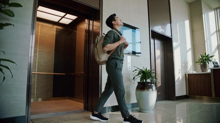 Stepping man in green shirt tightening tan backpack after elevator doors open, walking to reception