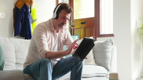 senior man using tablet with headphones, relaxing on couch at home