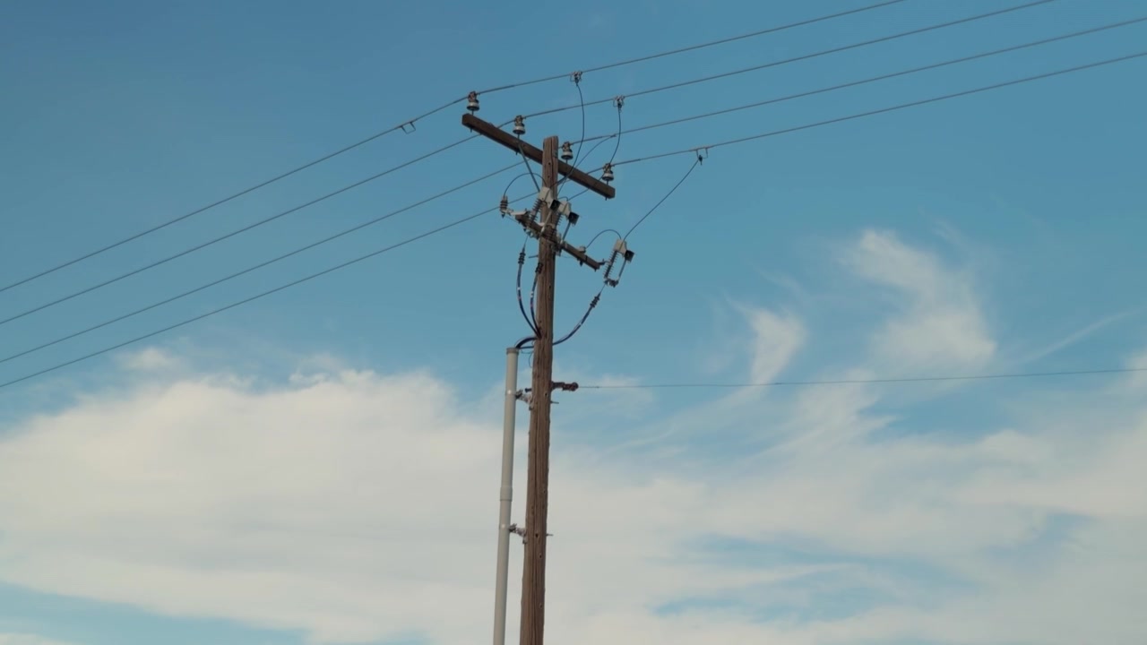 Steady downward pan along a wooden utility pole beneath power lines and clouds.