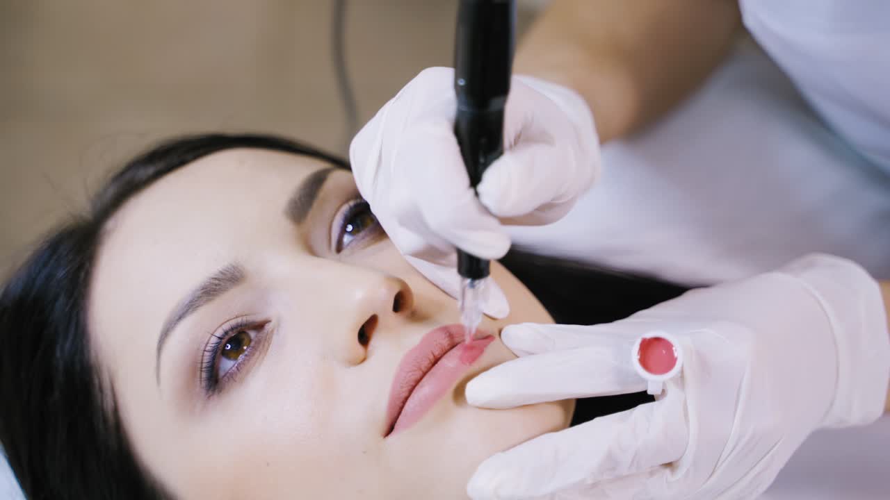 A beautician gently applies a pink hue pigment to the client's lips using a professional permanent makeup machine. Close-up.