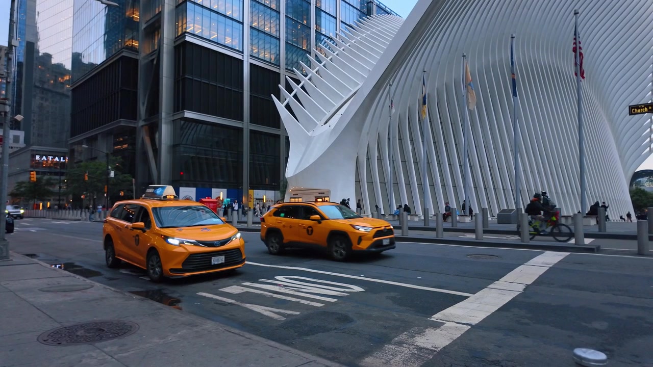 New York, USA, 5 June 2026 : Yellow taxi passing Oculus transit hub at dusk, lower Manhattan. Orange cab beside white ribbed structure, glass office tower and flags in fading light.