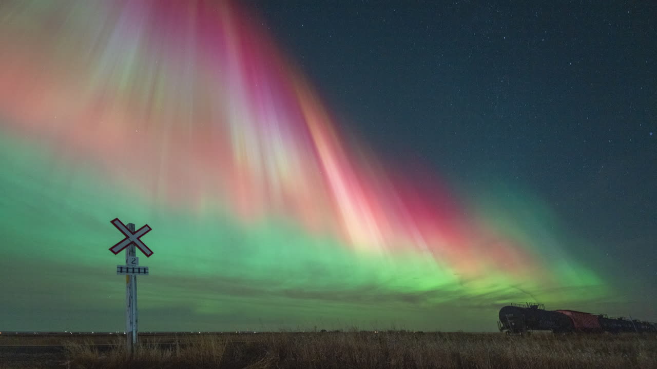 Colourful northern lights (Aurora Borealis) dance over parked train cars at a crossing in the prairie fields near Tuxford, Saskatchewan, Canada