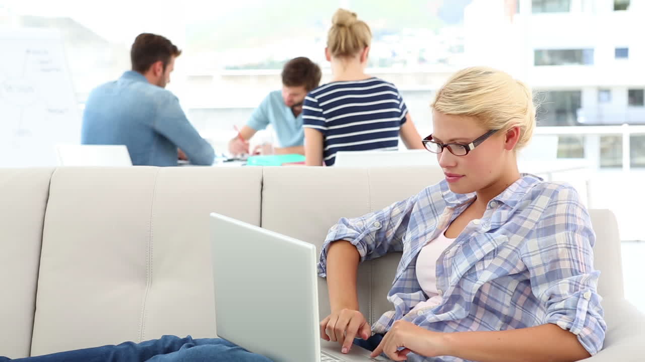Blonde woman using her laptop on sofa with colleagues behind