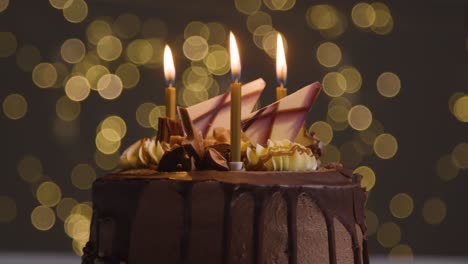 Studioaufnahme-Von-Dekoriertem-Schokoladengeburtstagskuchen-Mit-Brennenden-Kerzen,-Die-Vor-Bokeh-Hintergrundbeleuchtung-Ausgeblasen-Werden