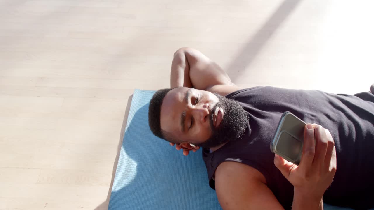 Uomo afroamericano felice che si rilassa sul tappetino usando lo smartphone a casa assolata, al rallentatore.