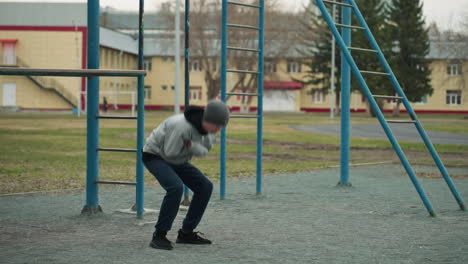 a young boy in a gray sweater and beanie performing a workout close to an iron pole with a blur view of people in the background