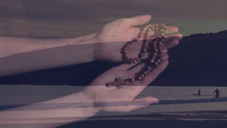 holding rosary beads, hands against serene beach landscape with distant figure