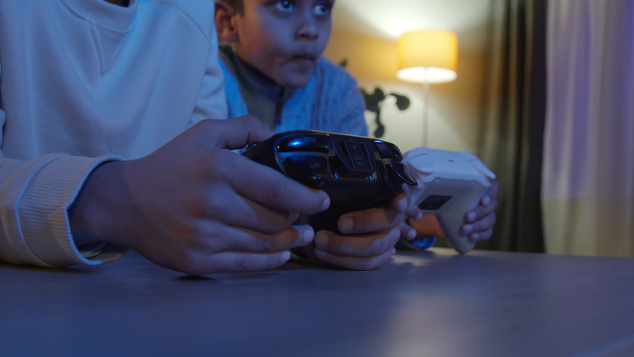 cerca de las manos de dos niños jóvenes en casa jugando con consola de juegos de computadora en la televisión sosteniendo controladores tarde en la noche 1