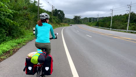 A-lone-female-cyclist-in-a-blue-t-shirt-riding-along-an-empty-roadway-in-the-Nan-Province,-Thailand