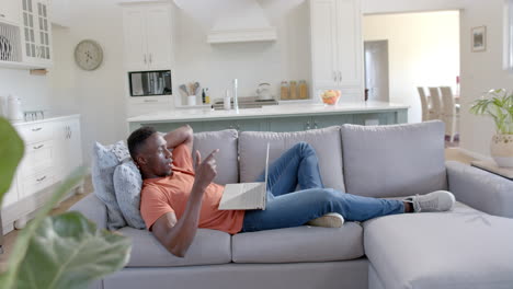 Happy-african-american-man-lying-and-having-video-call-on-laptop-in-sunny-living-room,-slow-motion