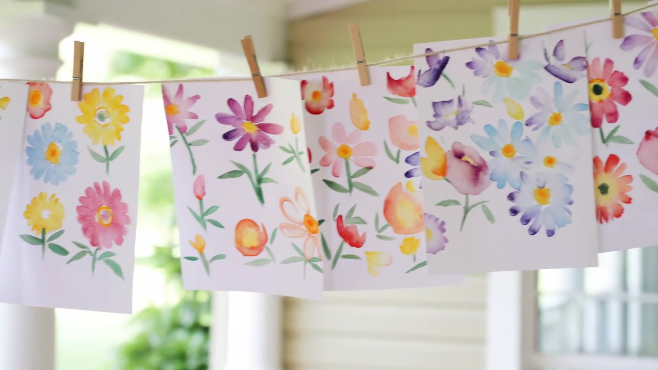 Række af håndmalede akvarelblomsterark hængende fra en snor på en overdækket veranda, fastgjort med træklemmer, blødt sollys og blide pastelfarver.