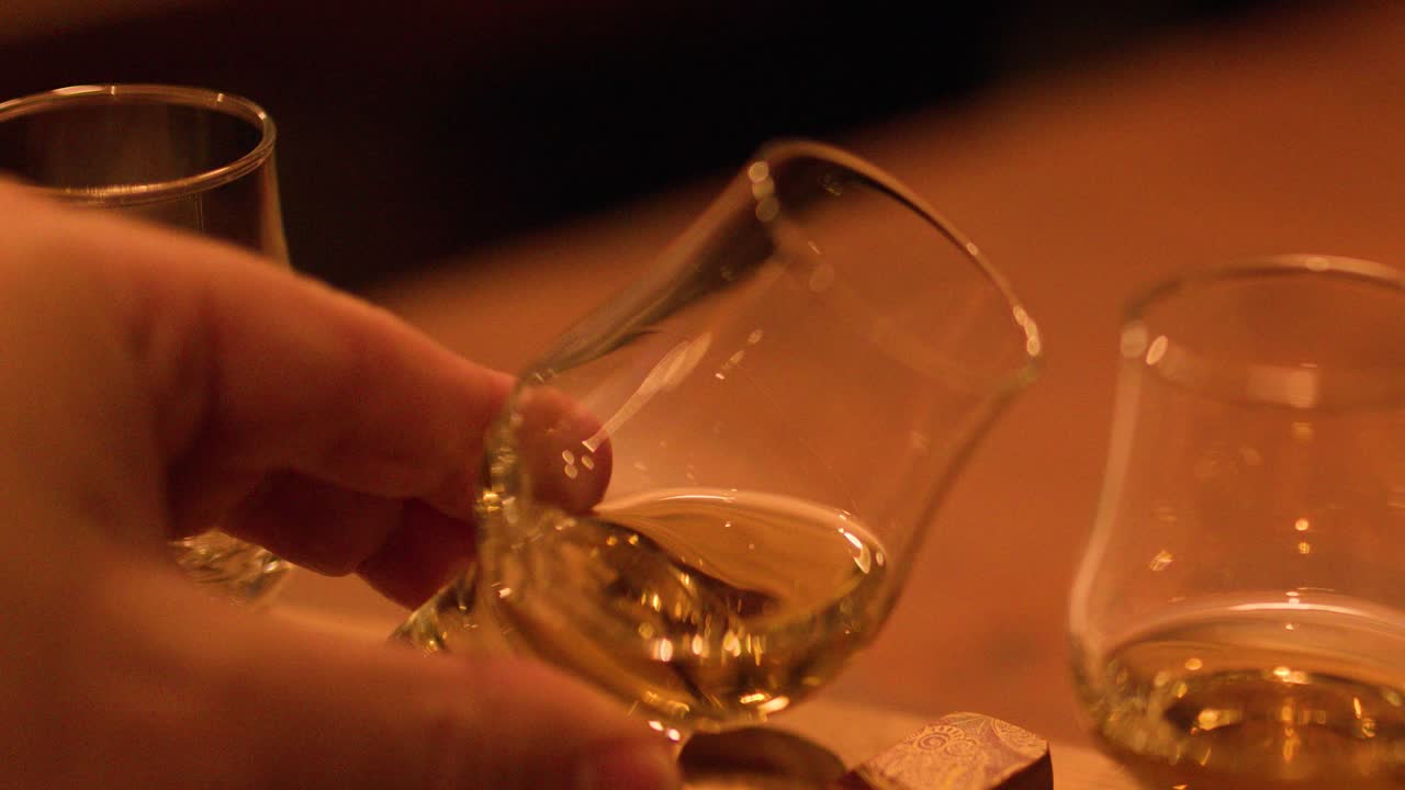Close-up of hand lifting whisky glass from tasting flight, warm ambient lighting, shallow focus