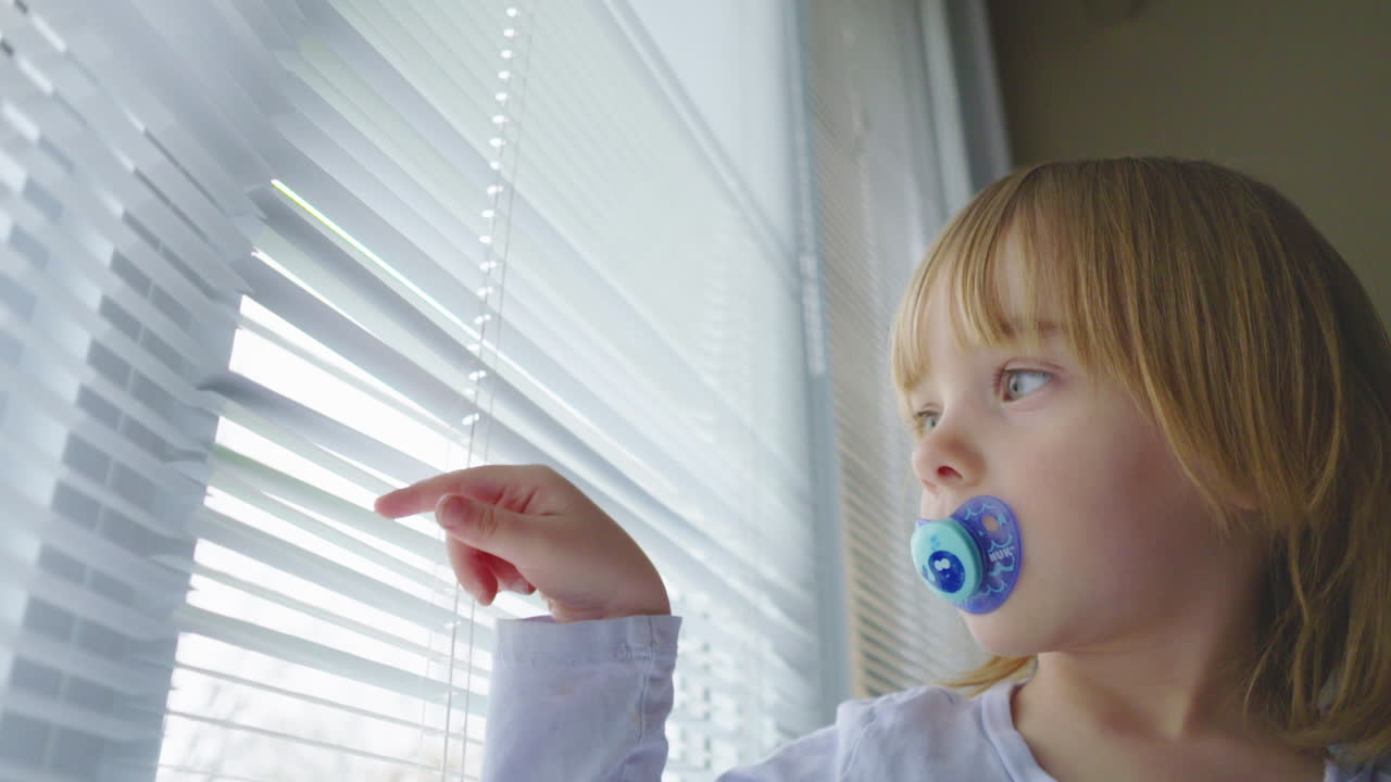A little girl with a pacifier in her mouth looks out a window and plays with the blinds