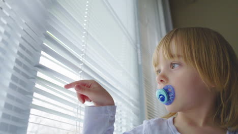 a little girl with a pacifier in her mouth looks out a window and plays with the blinds