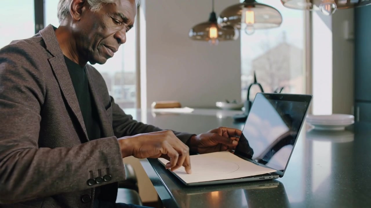 Sitting at kitchen bar, senior in dark blazer opening laptop, turning notebook pages starting work