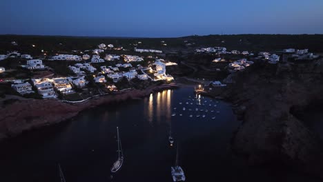 Vibrant-lights-of-Cala-Morell-reflect-onto-calm-bay-at-blue-hour-dusk,-aerial-pullback