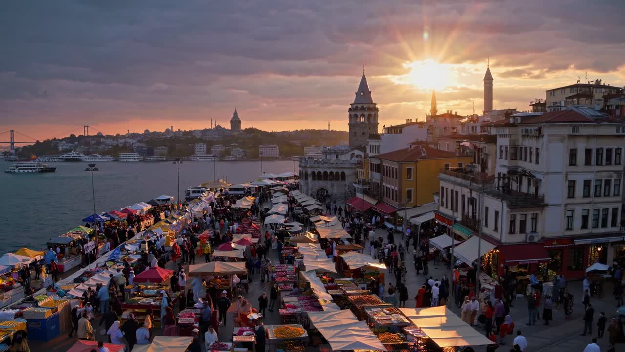 People celebrating Kurban Bayram by walking and shopping in a crowded market at sunset near Galata Tower in Istanbul, Turkey, with colorful tents and the Bosphorus in the background