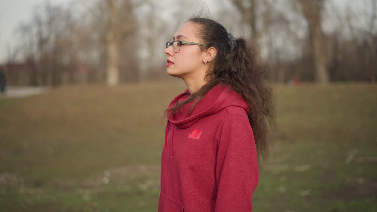 Asian Woman Walking In Park Back View Then Turning Head To Glance, Leash And Backpack Visible, Attentive Survey Of Surroundings, Casual Pace, Leafless Trees And Open Field Create Reflective Atmosphere