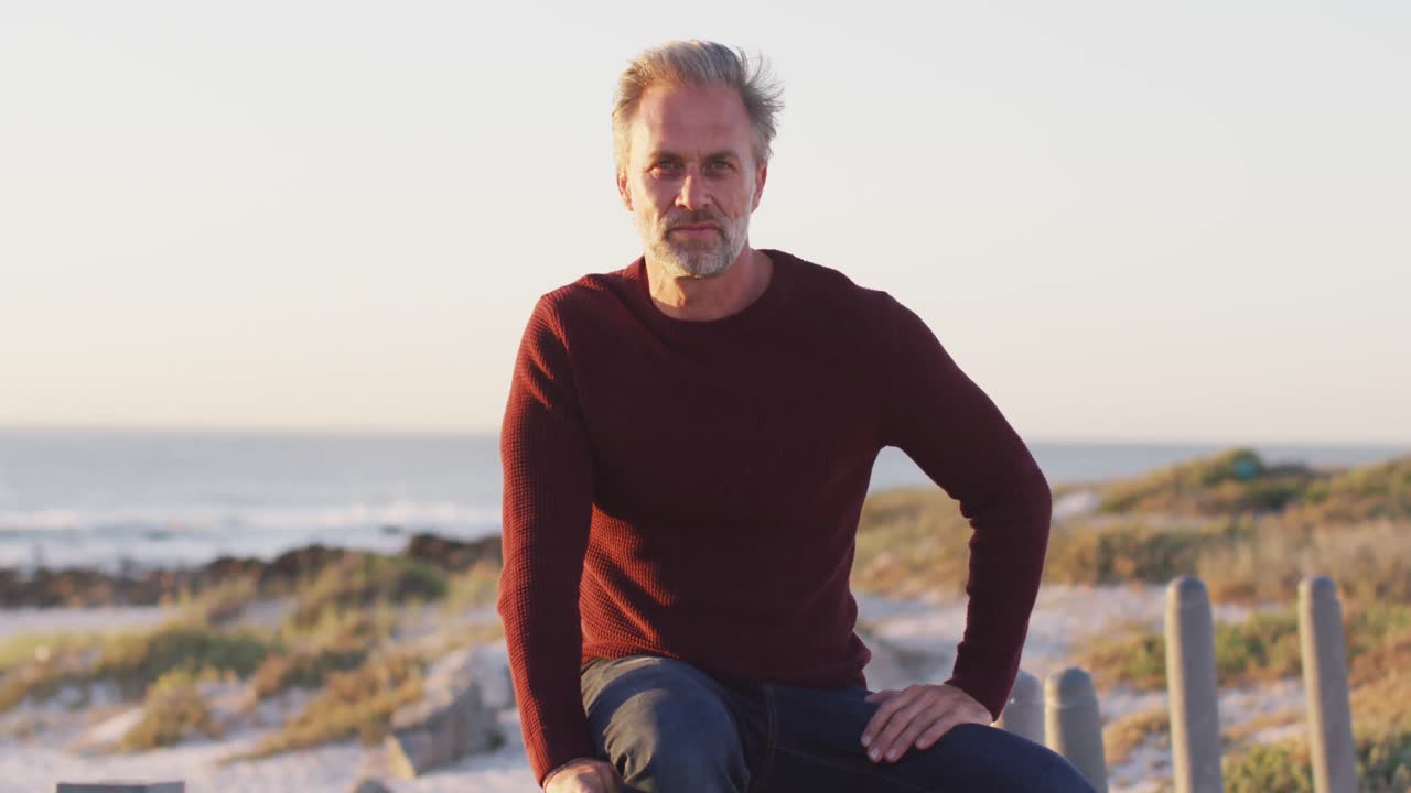 Portrait of happy caucasian man sitting, enjoying view and smiling on sunny day at the beach