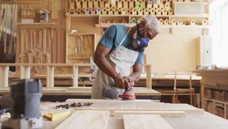 African-american-male-carpenter-wearing-protective-mask-sanding-a-wooden-plank-with-electric-sander