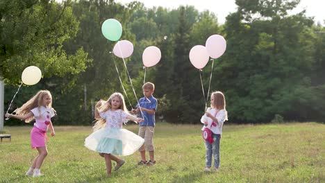 girls in dresses and with balloons in their hands are circling and jumping on the grass in the park. children play outdoors in summer. slow motion
