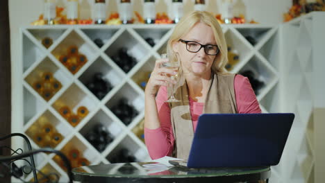 mujer catando vino sentada en una mesa en una bodega usando una laptop