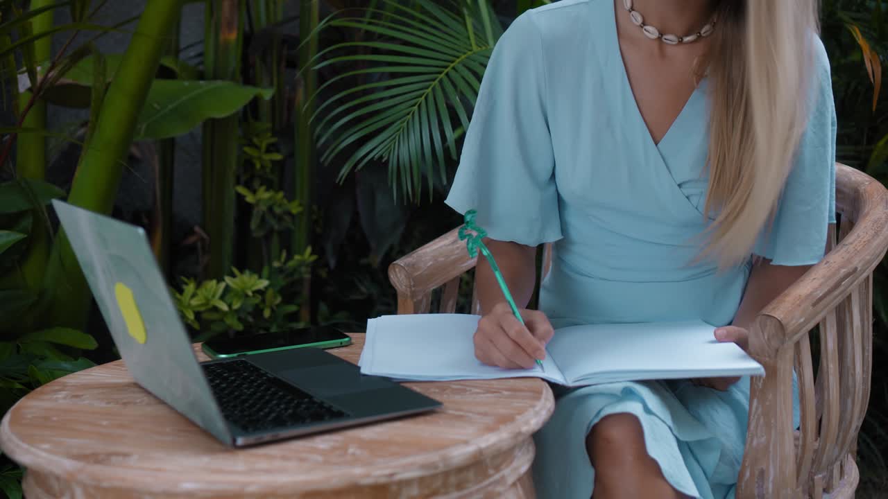 A young European girl in a blue dress remotely online working on laptop and looking into the screen on the backyard with green plants