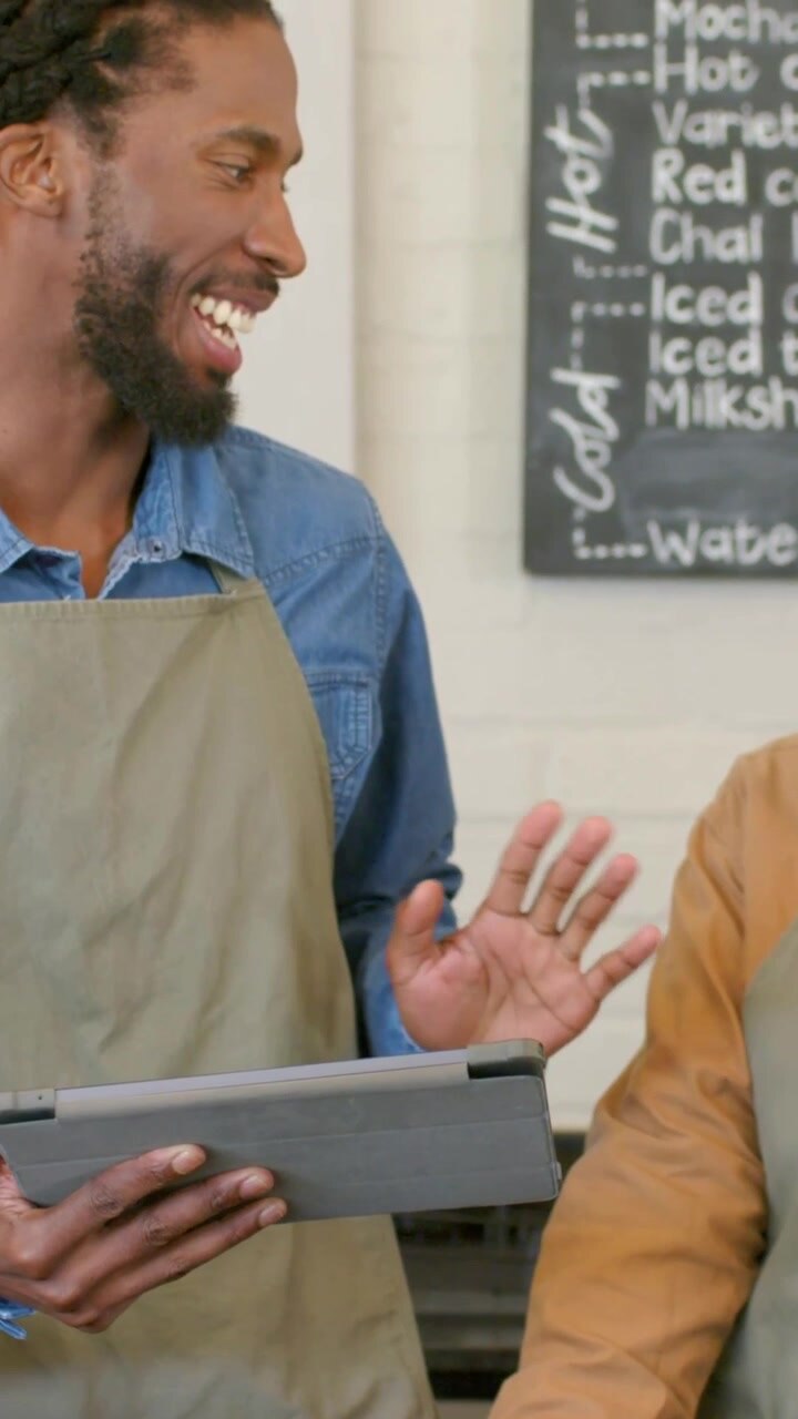 Turning woman barista smiling and talking at cafe counter, male barista holding tablet, gray aprons