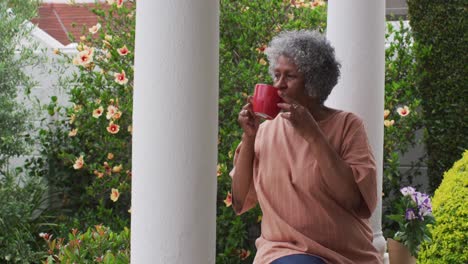 Ältere-Afroamerikanische-Frau-Trinkt-Kaffee,-Während-Sie-Auf-Der-Veranda-Des-Hauses-Sitzt