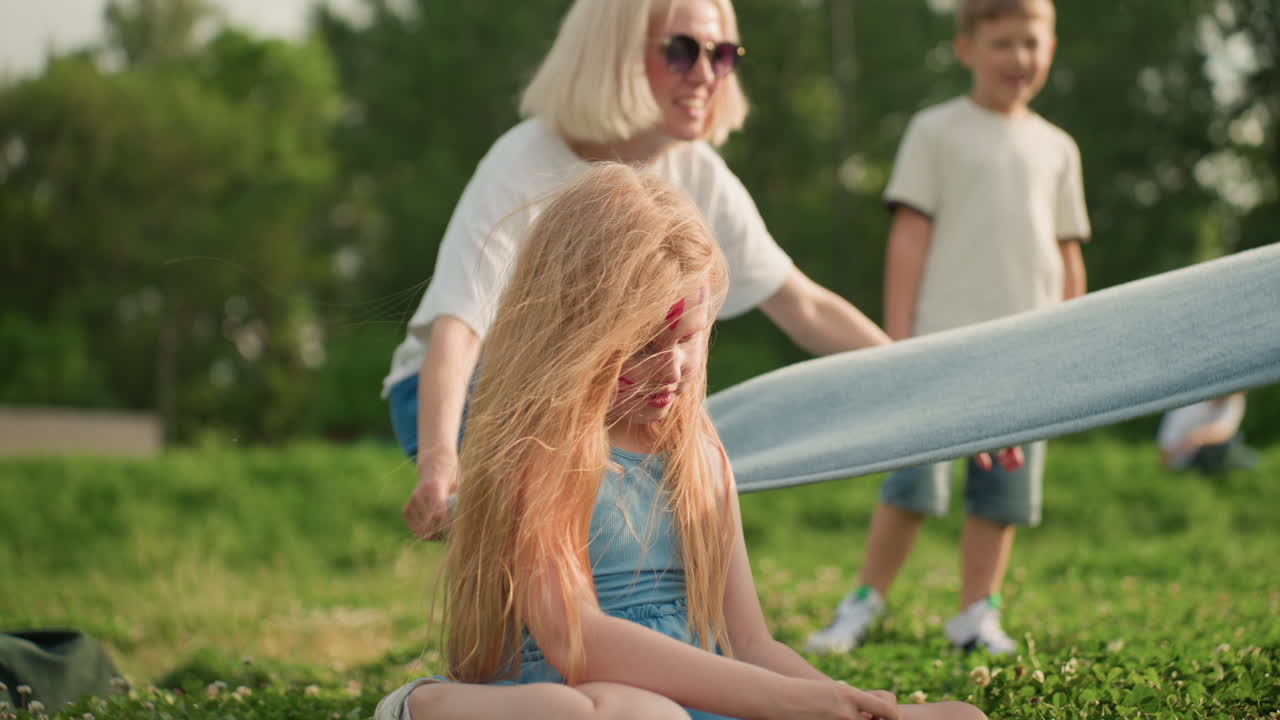 Face painted girl seated on grass while mother spreads picnic blanket nearby, sunlight filtering through trees, casual summer clothing, quiet moment before playful family gathering in park