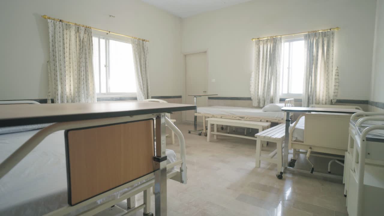 A vacant hospital room showing beds, chairs, and sunlight shining through windows.