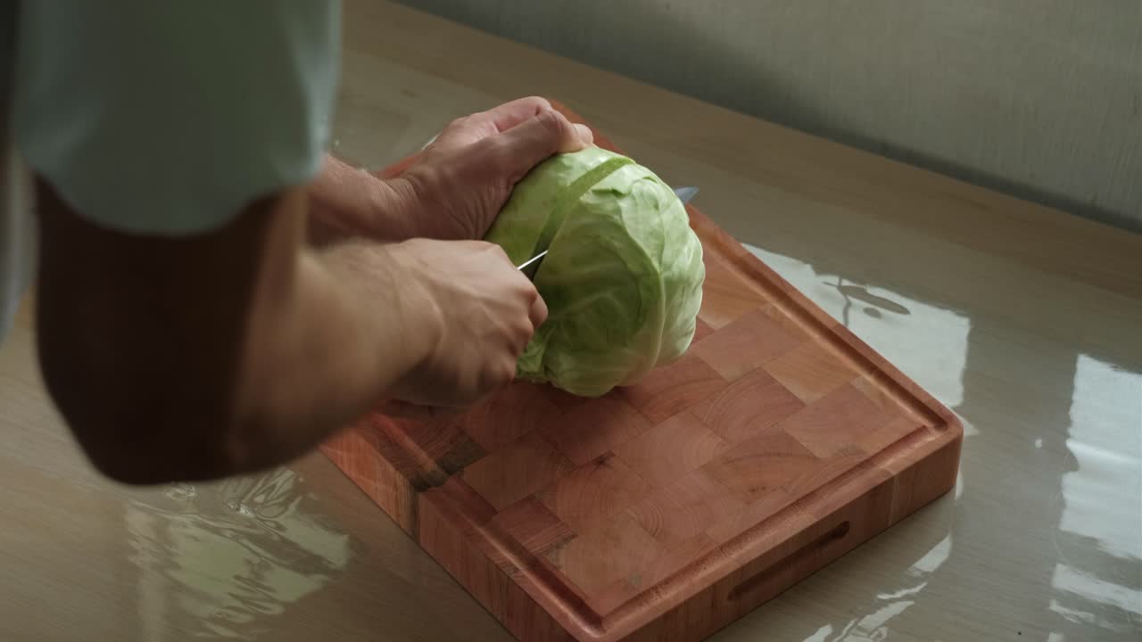 Young man slicing large cabbage in half with kitchen knife on wooden chopping board - close-up shot