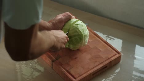 young man slicing large cabbage in half with kitchen knife on wooden chopping board - close-up shot
