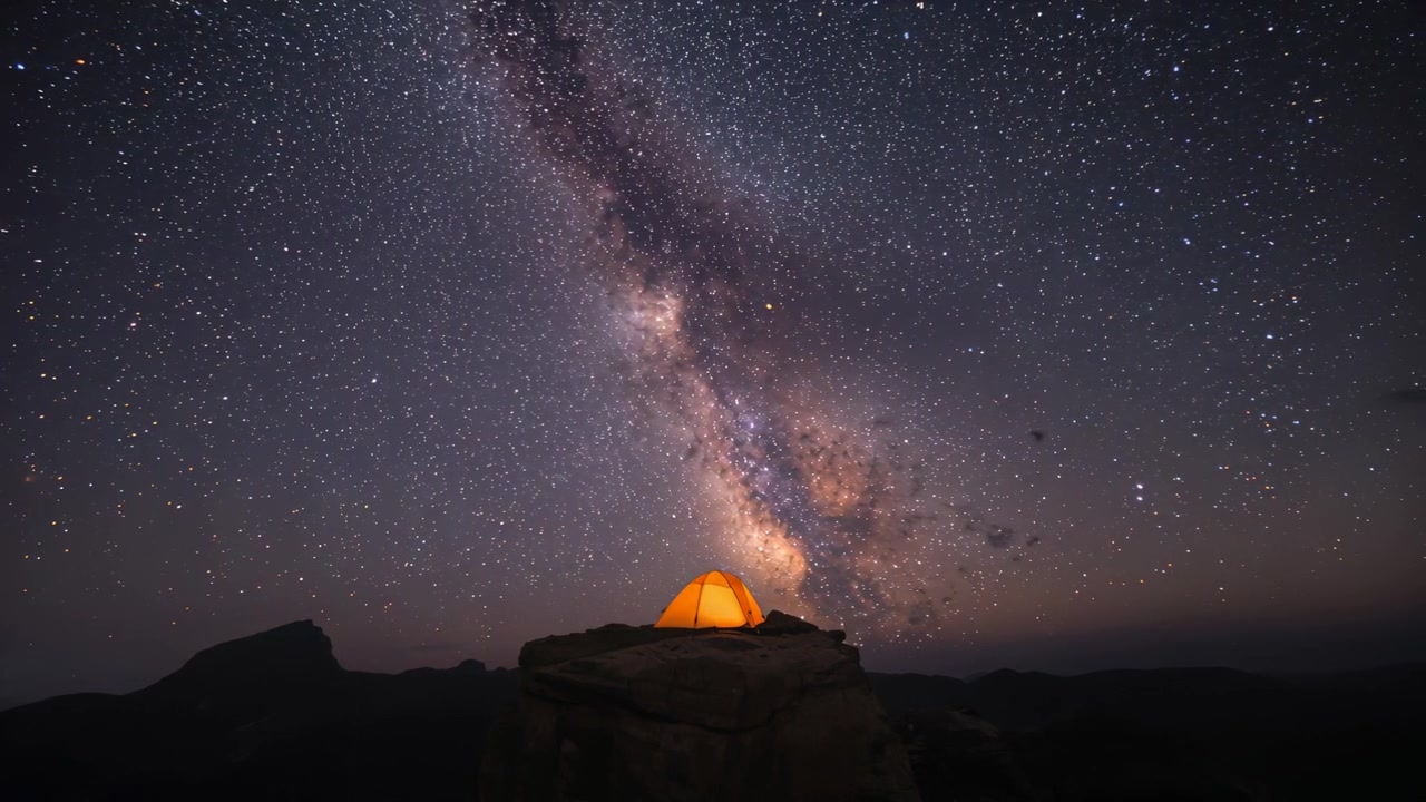 Camera starting timelapse capturing glowing orange tent on rock outcrop, recording Milky Way drift