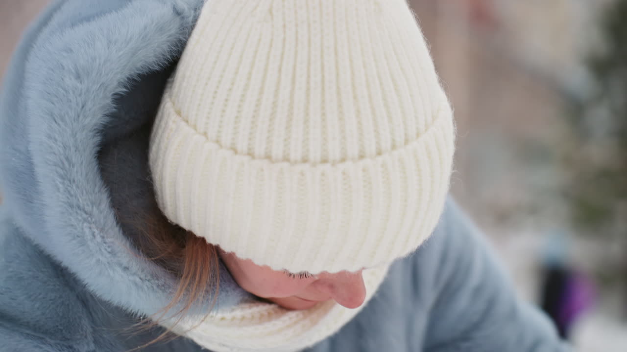 Wintermutter richtet ihre Mütze mit besorgtem Blick. Nahaufnahme-Porträt einer schützenden Mutter mit weißer Strickmütze, Schal und blauem Mantel, die den verschneiten Park nach ihrem Sohn absucht. Sichtbarer Atem in der kalten Luft, weiches Bokeh der Bäume.