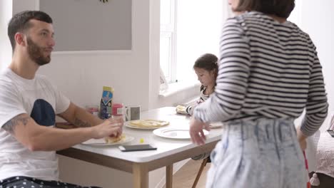 young family at home kitchen eating breakfast together with two daughters and talking