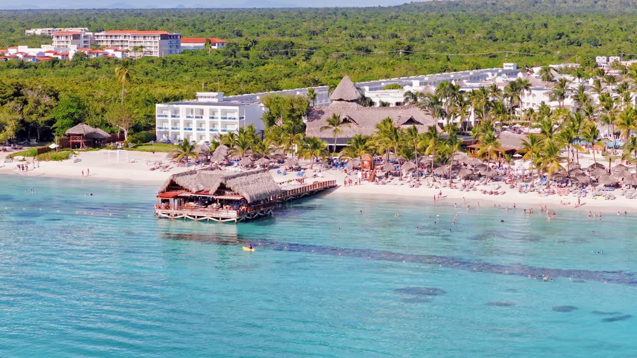 Restaurante en un muelle de madera en una playa tropical. Gente nadando en aguas turquesas en Bayahibe, República Dominicana. Vista aérea con dron