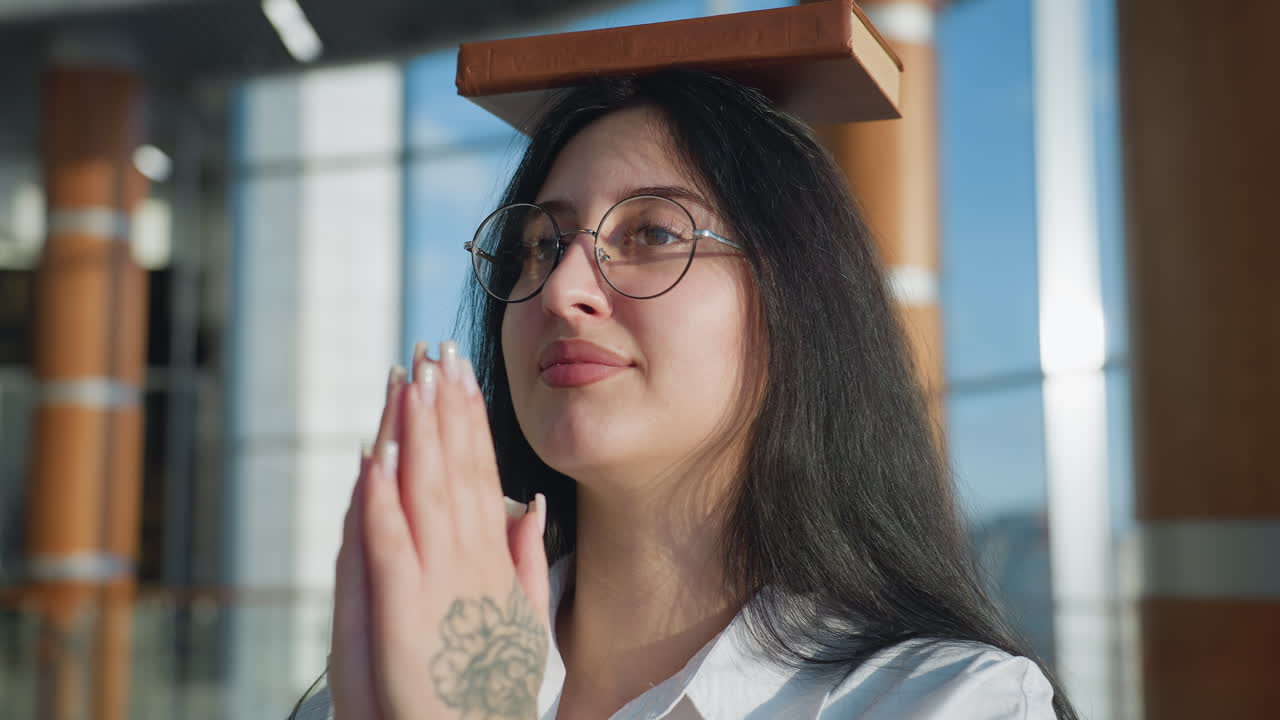 Close up of lady with glasses carefully walking while balancing book on head, hands joined in front showing floral tattoo, inside modern building with warm light and blurred architectural background