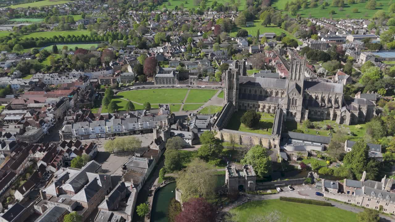 Rising aerial over Wells city where Gothic cathedral towers dominate a compact grid of medieval stone buildings and lawns within the green Somerset landscape